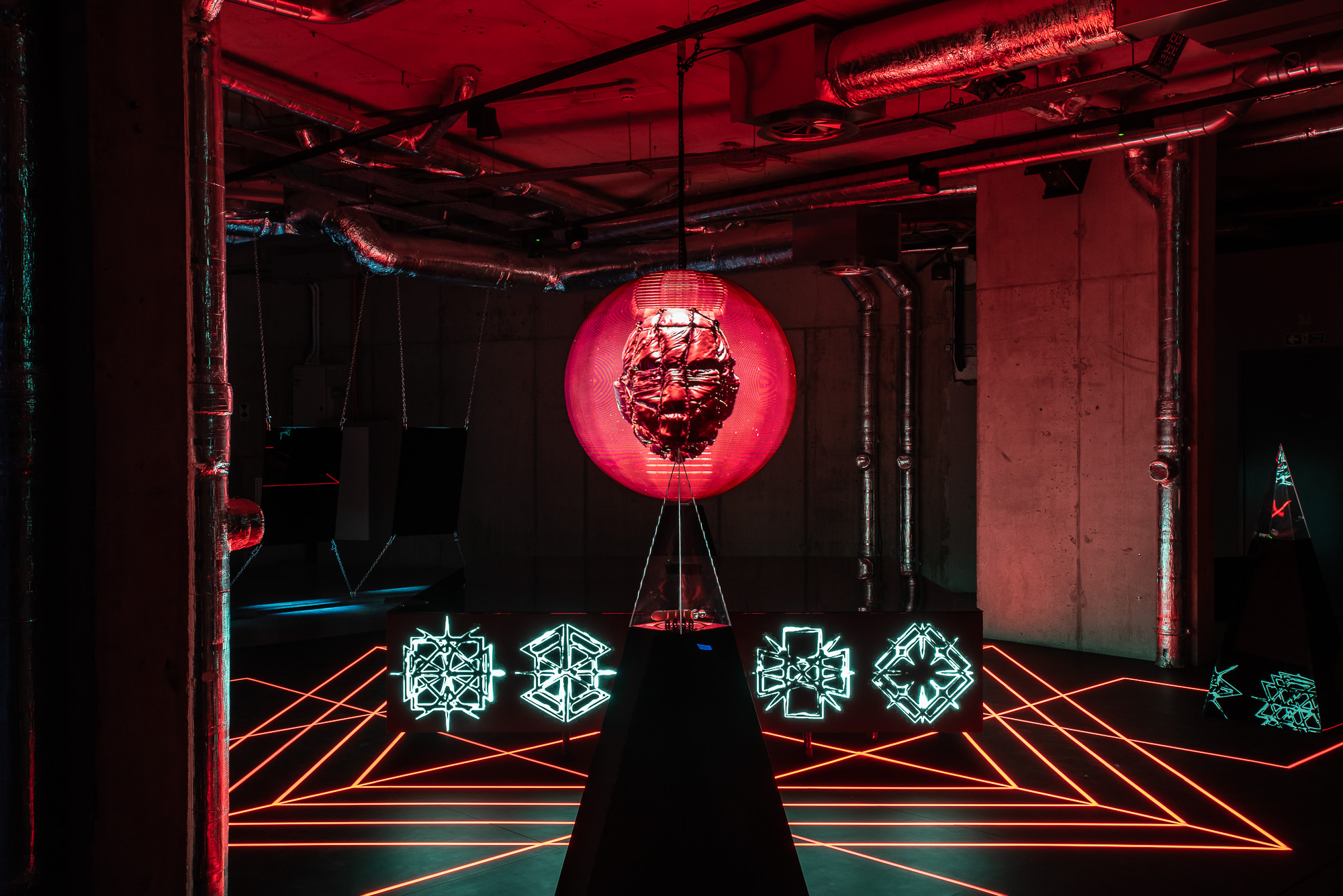 Installation view of dark room with red and blue lasers making up a geometric shape, with a sculpture of a face on top of a pillar. At the Arebyte gallery, London, UK (2023), photo by Max Colson